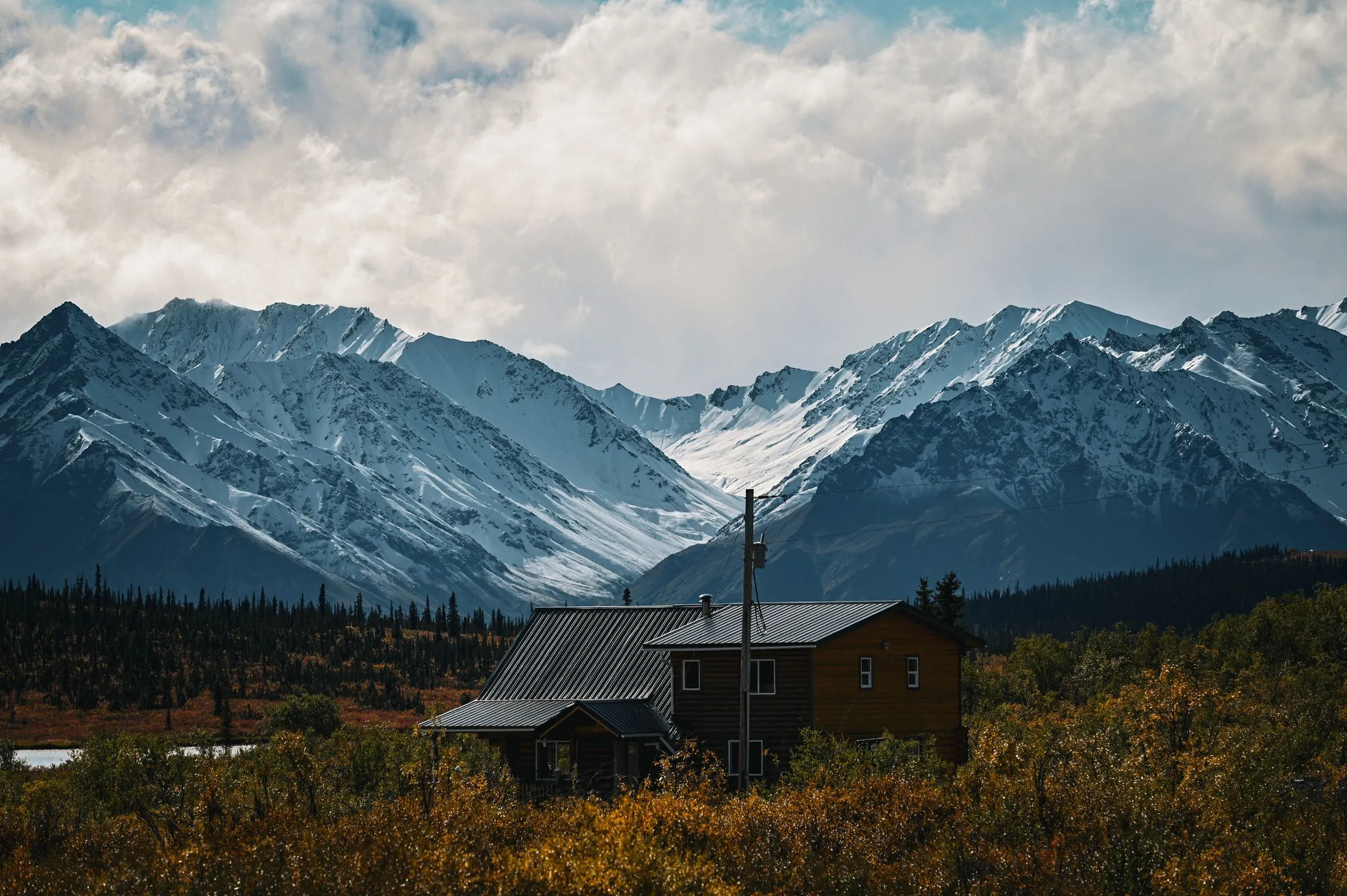 A small cabin with a metal roof below snowy mountains, with autumn colors in the foreground